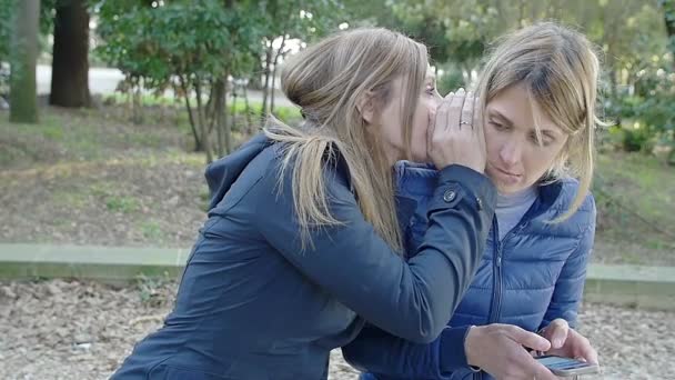 Две женщины веселятся в парке и делятся шокирующими новостями на ухо — стоковое видео