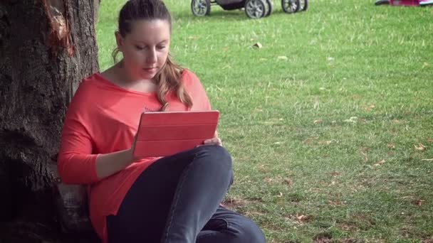 Mujer joven en el parque usando la tableta apoyada contra un árbol — Vídeo de stock