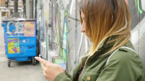 Mujer joven escribiendo mensajes de texto en el teléfono inteligente de pie junto a la pared de graffiti — Vídeo de stock