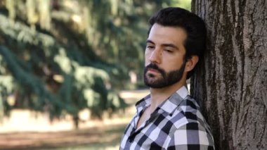 Genç endişeli adam düşünceli bir şekilde iç çekiyor parkta bir ağaca yaslanıyor.