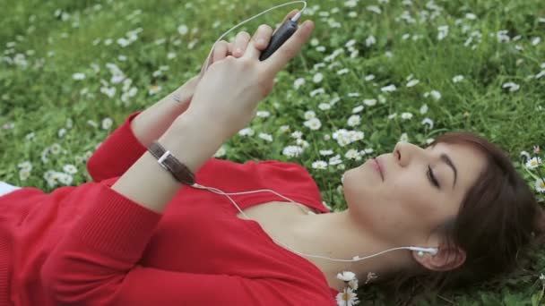 Hermosa mujer escuchar música mp3 desde el teléfono inteligente en el parque: hierba, al aire libre — Vídeo de stock