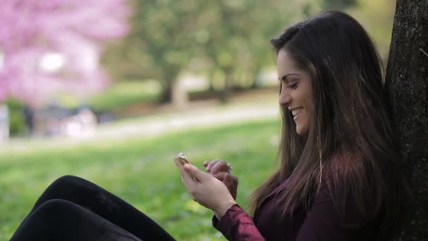 Doordachte jonge vrouw zitten in de buurt van boom gebruikt smartphone: glimlach, mobiele telefoon, — Stockvideo