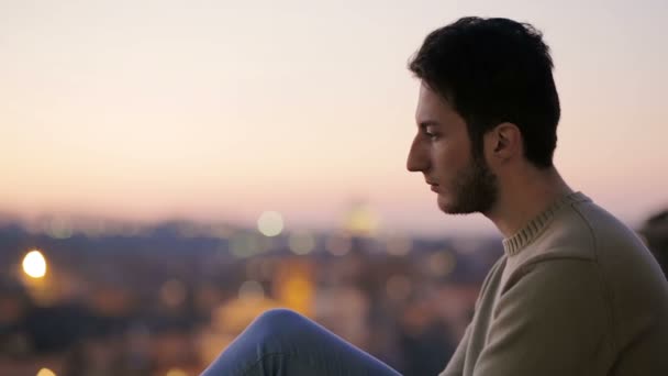 Jovem sentado, parece chateado. Homem em frente à paisagem ao pôr-do-sol — Vídeo de Stock