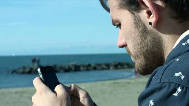 Ung man skriva SMS på pekskärmen av en mobiltelefon nära havet — Stockvideo