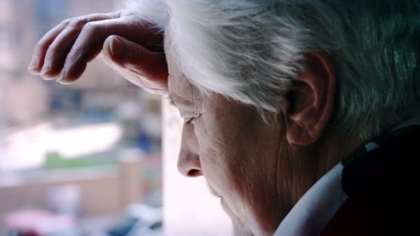 Worried old woman standing deep in her thoughts near the window — Stock Video