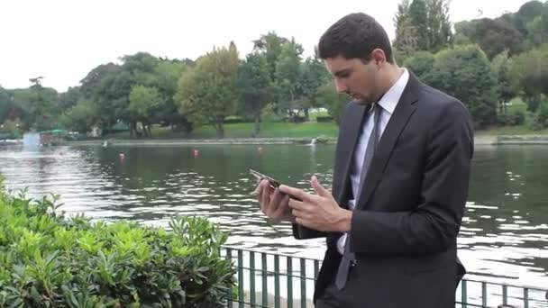 Young businessman using tablet pc near a lake — ストック動画