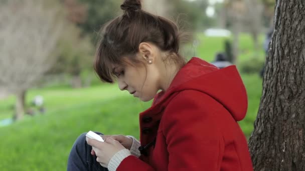 Aranyos lány használ smartphone a parkban: ülve, egyedül, boldog, üzenet — Stock videók