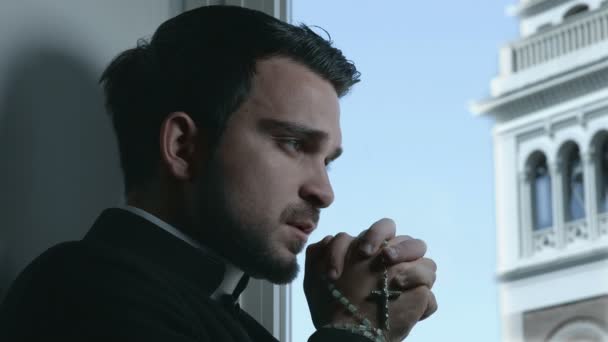 Priest prays with a rosary in his hand and bell tower in background — Stock Video