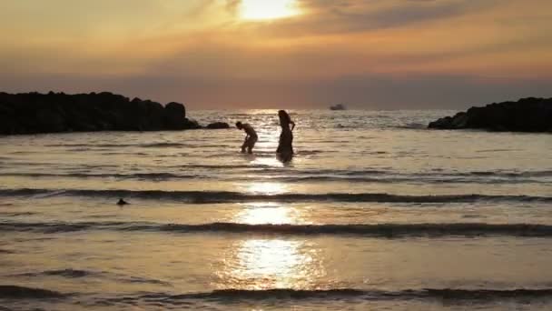 En pojke och flicka i havet vid solnedgången jakter och skämt med vatten — Stockvideo