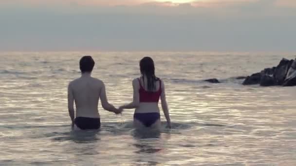 Iubitorii de mers pe jos în mare la apusul soarelui ținându-se de mâini și sărutându-se reciproc — Videoclip de stoc