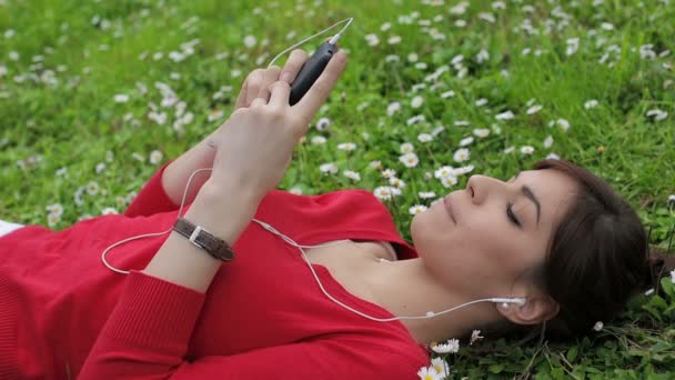 Roztomilá dívka pomocí smartphone v parku: poslech hudby, venkovní, ležící — Stock video