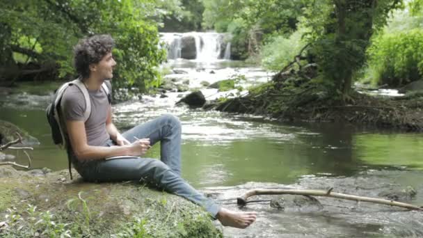 Jongen zit op de oever van de rivier — Stockvideo