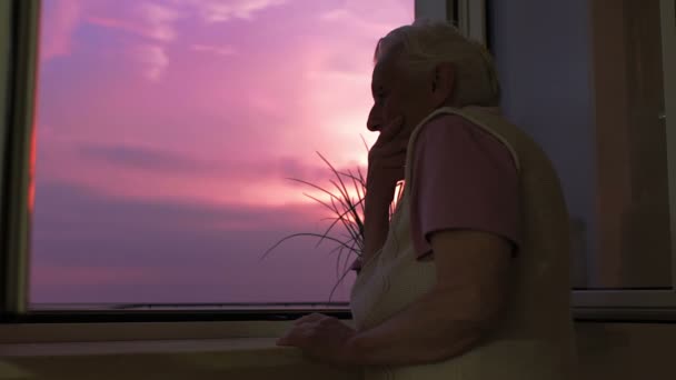 Nachdenklich - traurige alte Frau blickt aus dem Fenster auf den Sonnenuntergang — Stockvideo