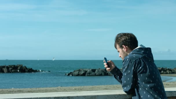 Молодий чоловік, використовуючи мобільний телефон, що спирається на стіну біля моря — стокове відео