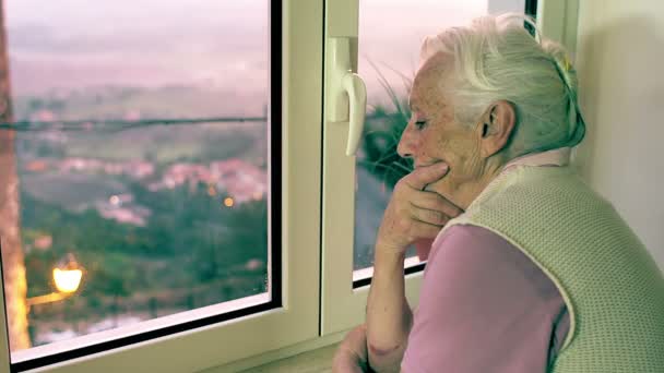 Nadenkend senior vrouw kijkt uit raam — Stockvideo