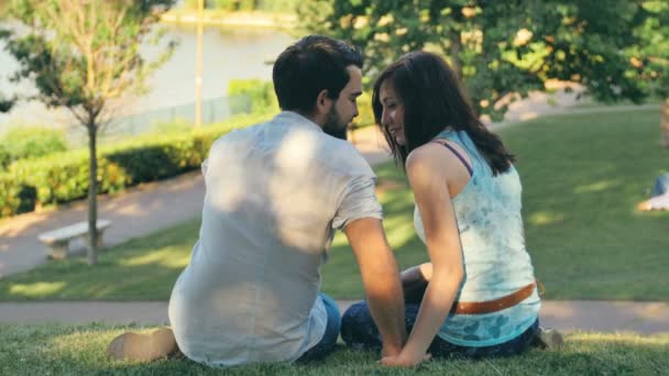 Two young people in love kissing sitting on the grass in a park — Stock Video