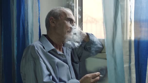 Старий сумний чоловік сидить біля вікна і курить вдумливо — стокове відео
