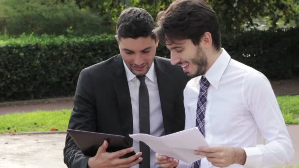 Два бизнесмена используют цифровые планшеты на скамейке в парке — стоковое видео