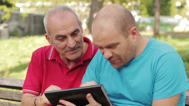 Äldre man med tablet Pc med hans vuxen son på park — Stockvideo