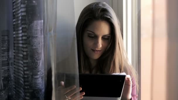 Молода жінка використовує планшет біля вікна: комп'ютер, навігація, веб, милий, стоячи — стокове відео
