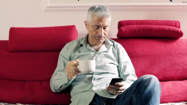 Милий чоловік п'є каву на дивані: фронтальна стрілянина з природним вікном — стокове відео