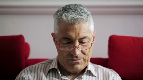 Homem de idade dos anos 50 tirando óculos sentados no sofá vermelho: sorriso, bonito, leitura — Vídeo de Stock