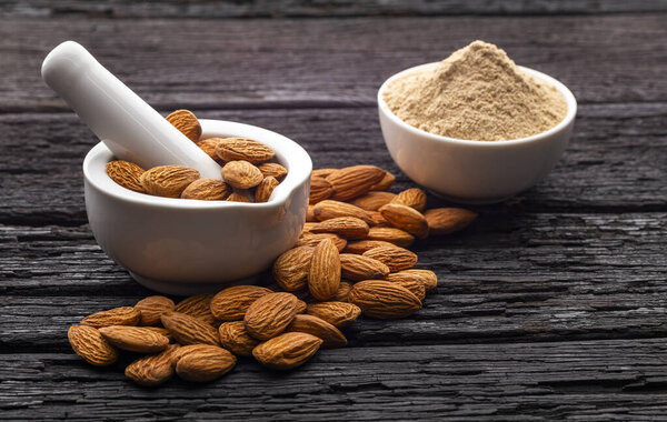 Natural almonds next to a white mortar and a bowl with almond flour on a rustic wooden surface.