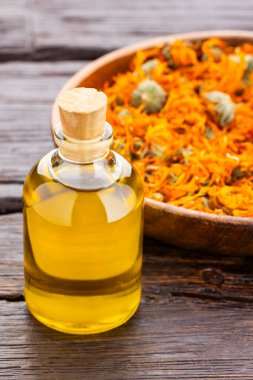 Kırsal bir masada bir şişe Calendula yağı, yanında da ahşap bir kasede kurumuş taç yaprakları.