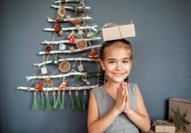 Mutlu kız, orijinal alternatif Noel ağacının yanında, donmuş dallarla yapılmış, küçük ekolojik oyuncaklar ve kağıt çelenklerle süslenmiş, sıfır atık ve plastiksiz tatillerle dolu bir hediye kutusu tutuyor.