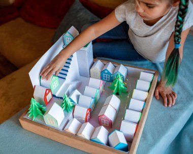 Origami takvimi, Noel köyü, kağıt işleri. Şirin bir kız, içinde sayılar ve kağıt yeşili ağaçlar olan küçük kağıt evlere bakıyor, çocuklarla mevsimsel aktiviteler, kapalı bir yaşam tarzı.