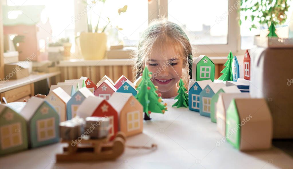 Origami advent calendar, Christmas village, paper craft. Cute girl looking upon tiny paper houses with number and paper green tree, seasonal activity with kids, indoor lifestyle