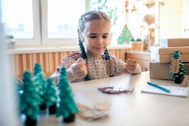Şirin bir kız Noel için ailelere Noel hediyesi hazırlıyor ve onlara etiket imzalıyor. Küçük bir çam ağacı, etiket ve bayram kutuları gibi yeşil bir koni. El yapımı hediyeler, sıfır atık tatil.