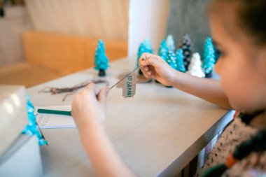 Şirin bir kız Noel için ailelere Noel hediyesi hazırlıyor ve onlara etiket imzalıyor. Küçük bir çam ağacı, etiket ve bayram kutuları gibi yeşil bir koni. El yapımı hediyeler, sıfır atık tatil.