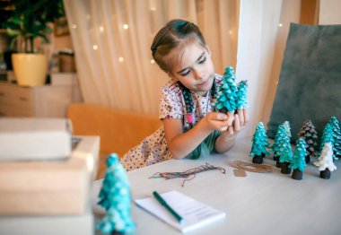 Şirin bir kız Noel için ailelere Noel hediyesi hazırlıyor ve onlara etiket imzalıyor. Küçük bir çam ağacı, etiket ve bayram kutuları gibi yeşil bir koni. El yapımı hediyeler, sıfır atık tatil.