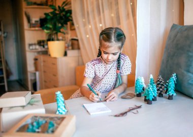 Şirin bir kız Noel için ailelere Noel hediyesi hazırlıyor ve onlara etiket imzalıyor. Küçük bir çam ağacı, etiket ve bayram kutuları gibi yeşil bir koni. El yapımı hediyeler, sıfır atık tatil.