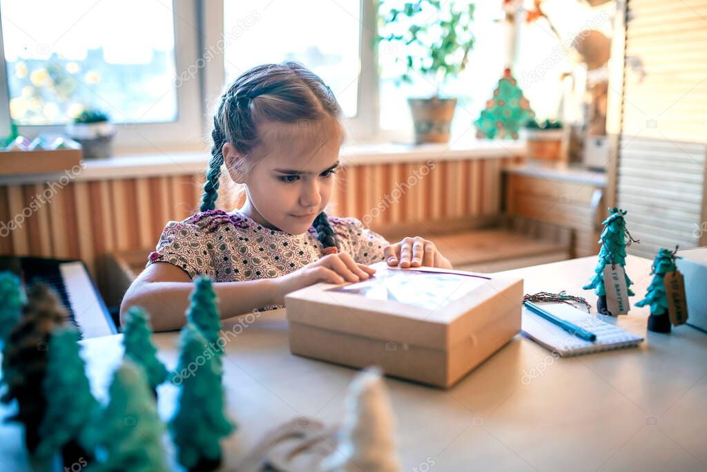 Cute girl preparing DIY gifts and signing tags to parents and family for Christmas, a green cone as if it is small toy pine tree, tags and festive boxes, handmade presents, zero waste holidays