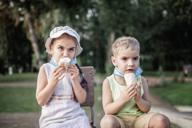 Yüzü maskeli şirin bir kardeş karantina sırasında dondurma yiyor, açık hava yakın plan portresi, yeni normal gerçeklikte sıcak hava.