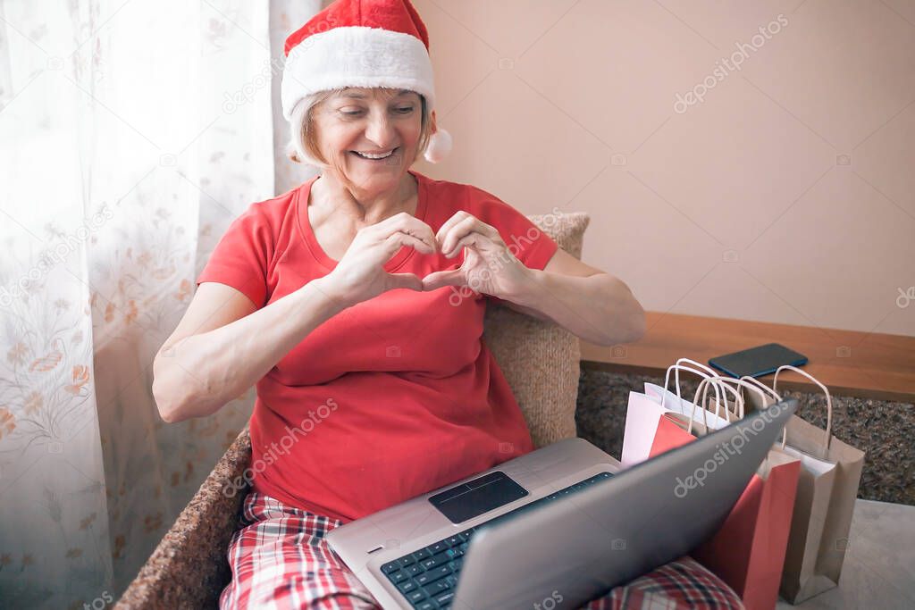 Safe online Christmas celebration. Senior woman in Santa red hat giving present to her family virtually with internet and notebook. Video call. Stay home, distant holiday, emotional indoor lifestyle