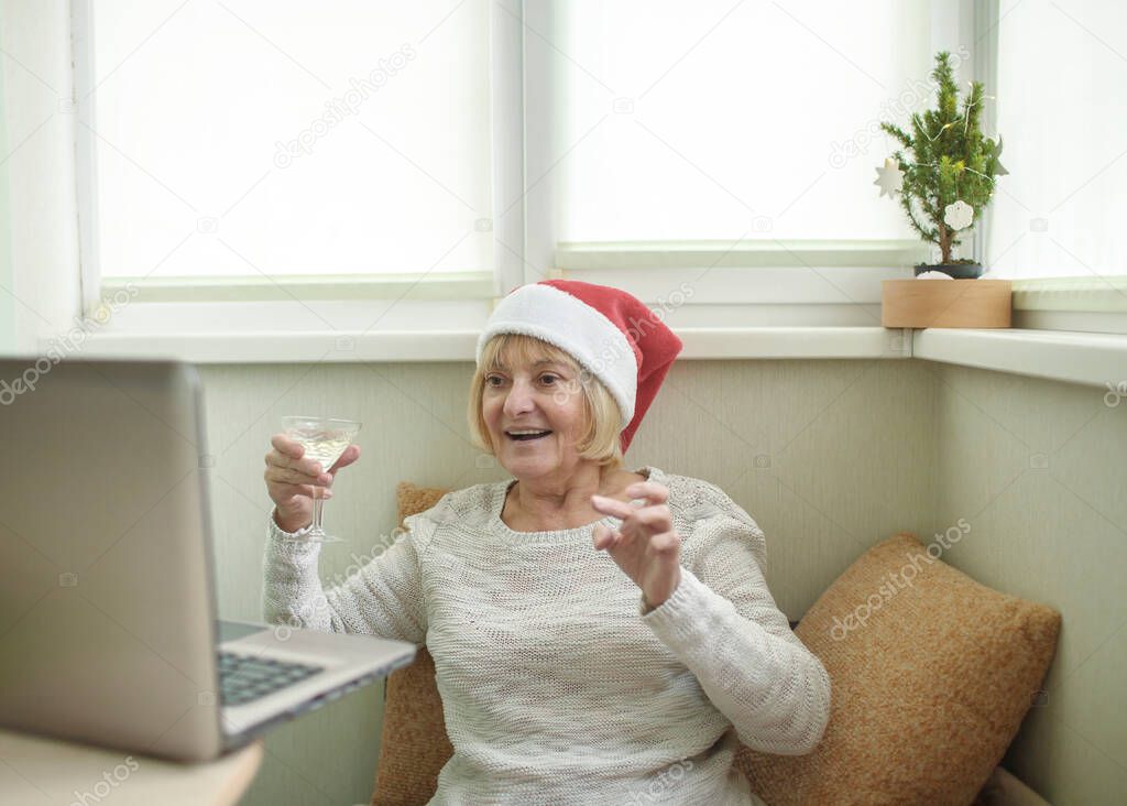 Safe online celebration. Senior woman holding a glass of wine and celebrating Christmas with her family virtually via internet and notebook at home. Video call. Distant holiday, indoor lifestyle