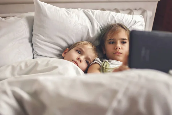 Двое братьев и сестер смотрят видео в мобильном электронном устройстве в кровати, гаджетная зависимость, образ жизни — стоковое фото