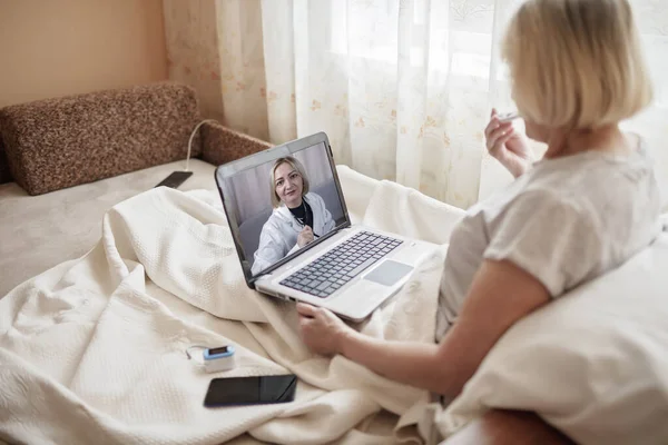 Old woman in bed looking at screen of laptop and consulting with a doctor online at home, telehealth