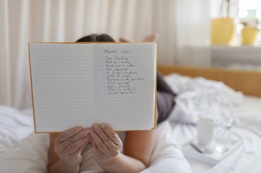 Akıl sağlığı, olumlu düşünme ve sağIık konsepti. Çekici kadın yatakta uzanıyor, günlükle duygu, bir bardak su ve minnet listesi hakkında stres atma alıştırması yapıyor.
