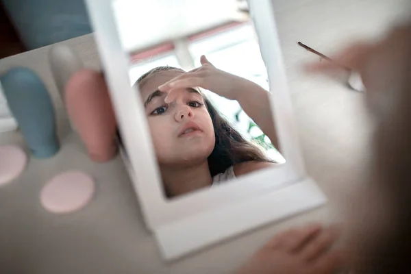 Девочка-подросток с длинными волосами наносит маску для лица перед зеркалом, салон красоты дома — стоковое фото