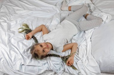 Awaking girl in white clothes with two braids lying on bed in bedroom in the morning, sleep care clipart