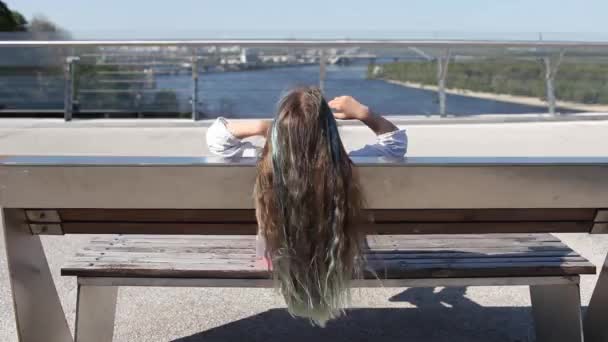 Brunette preteen girl with blue color highlights sitting on bench on bridge, θέα πίσω, καλοκαιρινή μέρα — Αρχείο Βίντεο