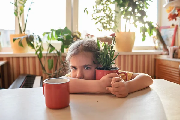 Girl drinking coffee and enjoying green blooming flower replanted in red mug, home floral decor — стоковое фото