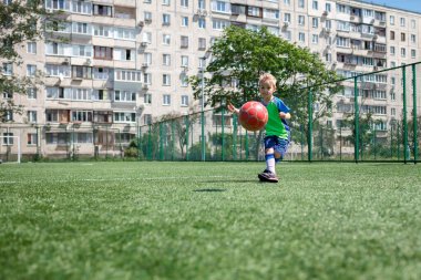 Bahçede futbol oynayan mavi ve yeşil formalı küçük bir çocuk, genç bir futbolcu.