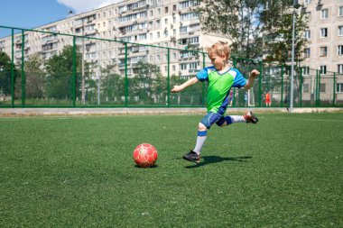 Bahçede futbol oynayan mavi ve yeşil formalı küçük bir çocuk, genç bir futbolcu.