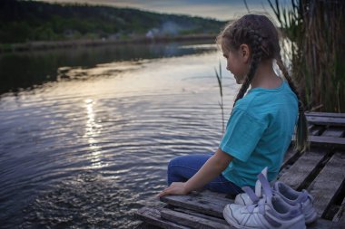 Göl kıyısındaki rıhtımda bacaklarıyla su sıçratan neşeli kız, mutlu bir çocukluk.