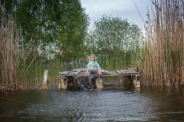 Joyful girl splashing with legs in water on the pier at lakeshore, happy childhood — Stock Photo, Image